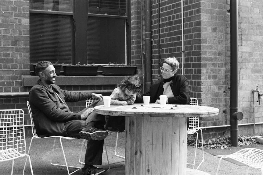 A black-and-white image of a smiling, dark-haired man, a little girl and a woman sitting at a table in a courtyard.