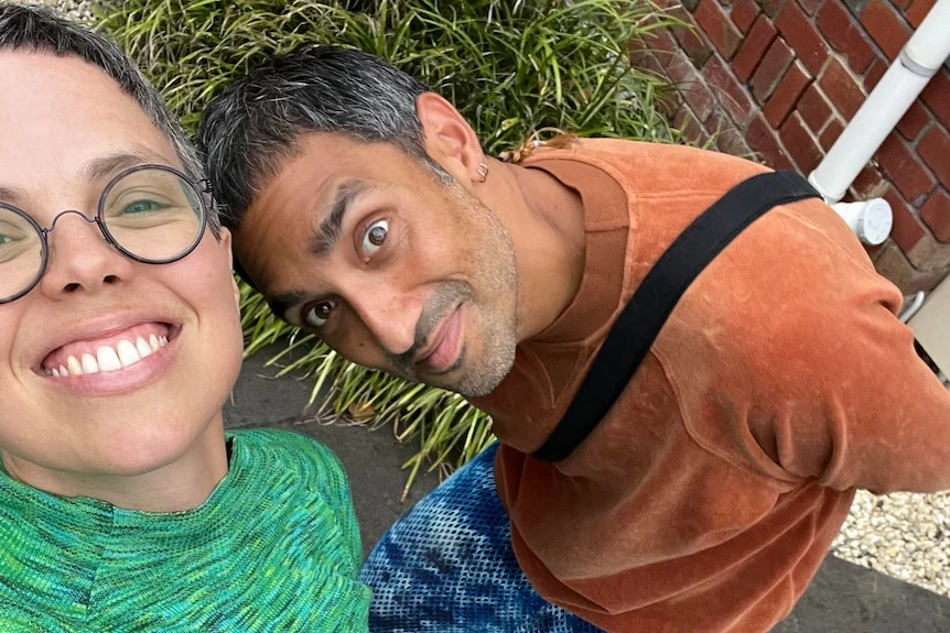 A smiling woman with short hair and glasses takes a selfie with a dark-haired man near a brick building.
