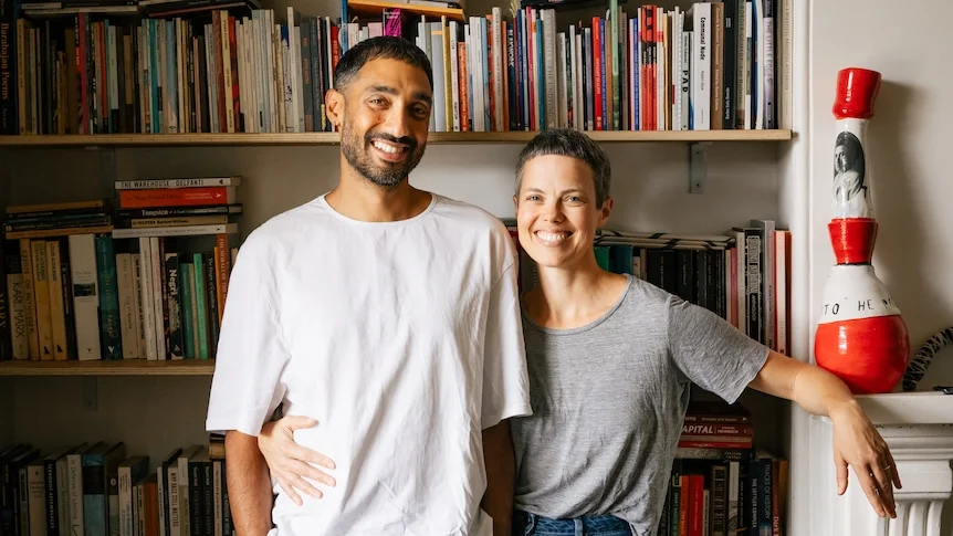 A smiling dark-haired man and a woman with short, dark hair standing in front of bookshelves in a room.