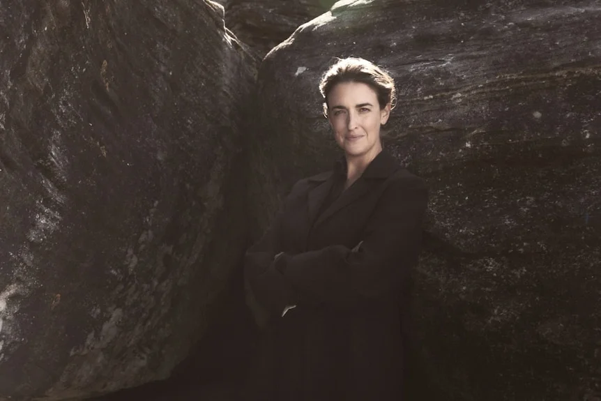 Lucianne Tonti; wearing an oversized black suit jacket, standing in front of large rocks, the sun glinting off her hair.