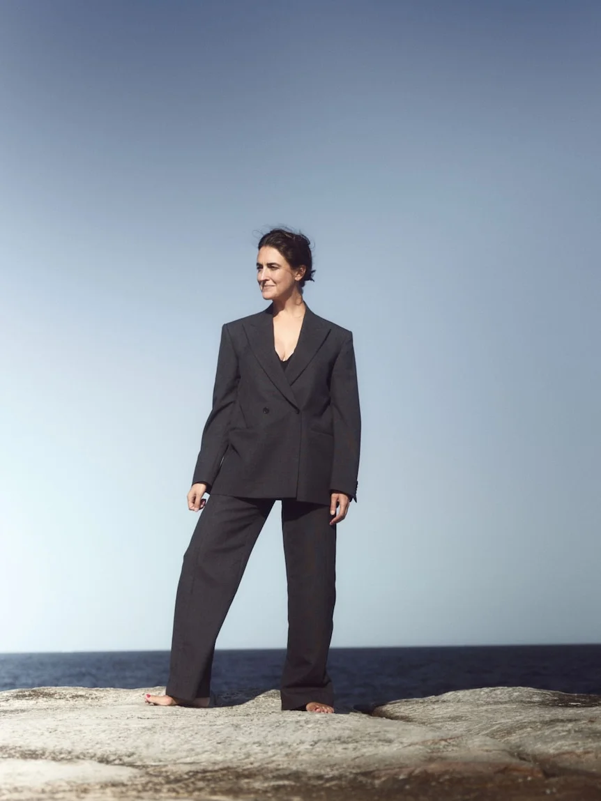 Lucianne Tonti; a woman wearing an oversized black suit, standing on rocks by the sea. A blue sky is behind her.
