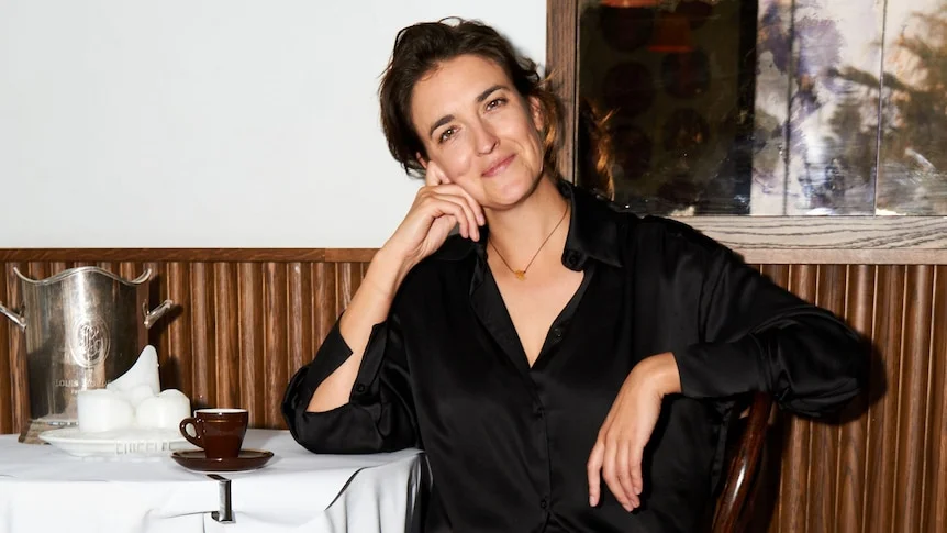 Lucianne Tonti; a woman with long dark hair sitting in a restaurant, her elbow on the table and her head resting on her fist.