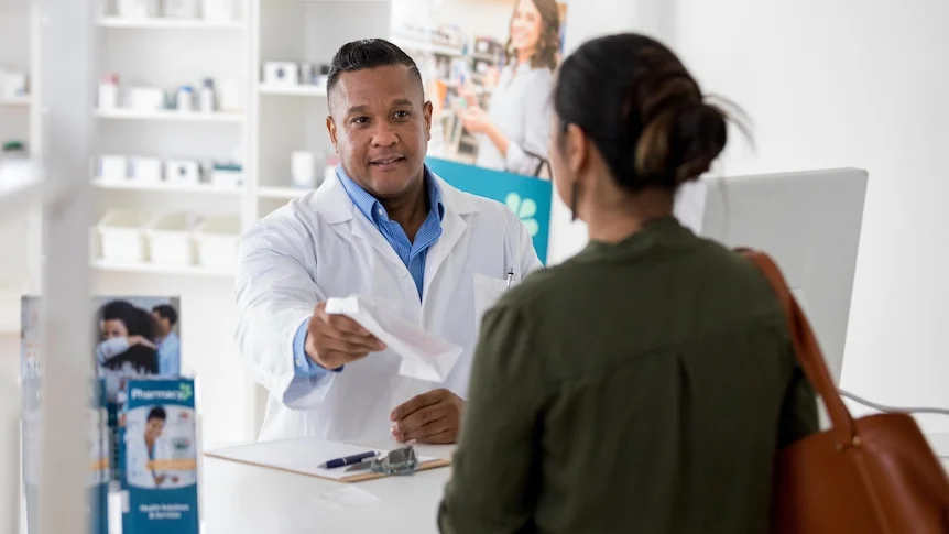 A male pharmacist hands a box of medications to a female patient. They are smiling.