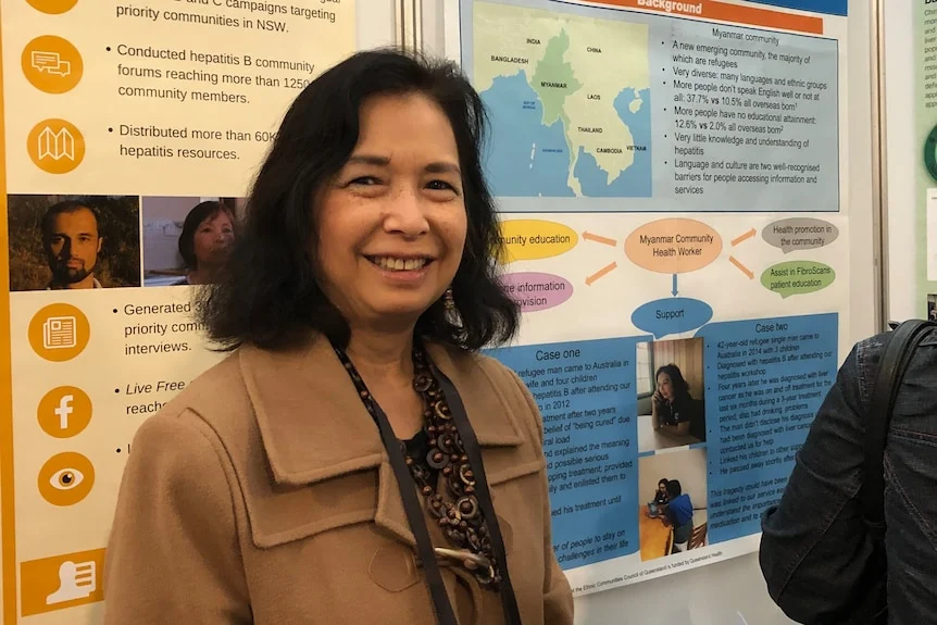 Evelyn, woman with short dark hair smiles in front of an information board about hepatitis and Myanmar.