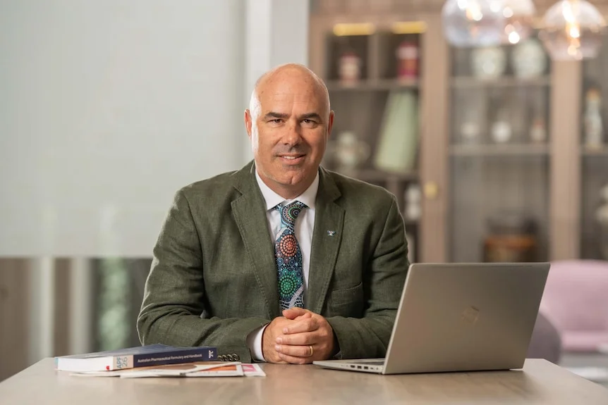 Mark is wearing a brown suit sits at a desk with books and an open laptop. He smiles at the camera.