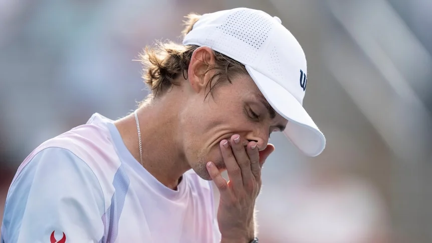 Alex de Minaur rubs his face with his left hand while wearing white cap