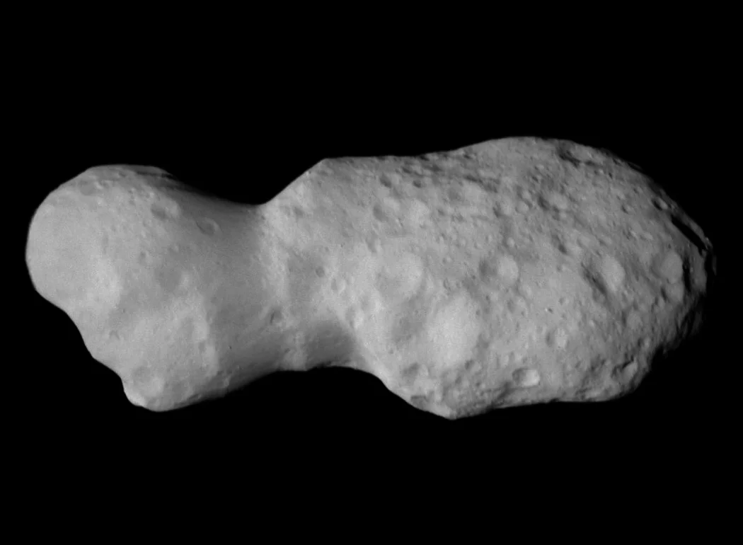 A gray, irregularly shaped asteroid with a lumpy surface and numerous impact craters, reminiscent of a contact trinary or dirty snowball planet form, set against a black background.