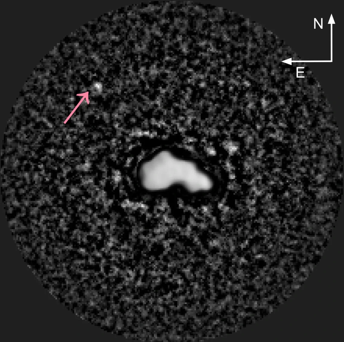 Black and white circular astronomical image with a bright central area and a red arrow pointing to a faint spot; labeled compass shows north and east directions. The faint feature indicated by the arrow may represent an early stage of dirty snowball planet form, potentially providing insight into contact trinary evolution.
