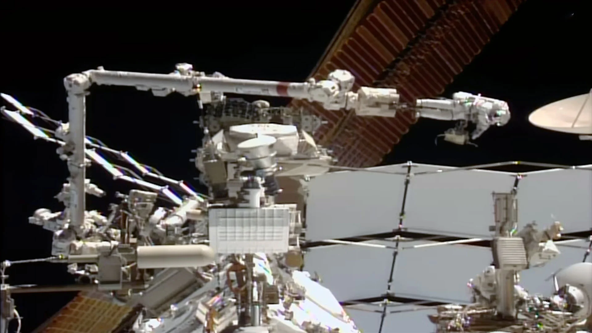 an astronaut wearing a white spacesuit appears to be sideways (in relation to the camera) as he is moved by a large robotic arm outside of a space station.