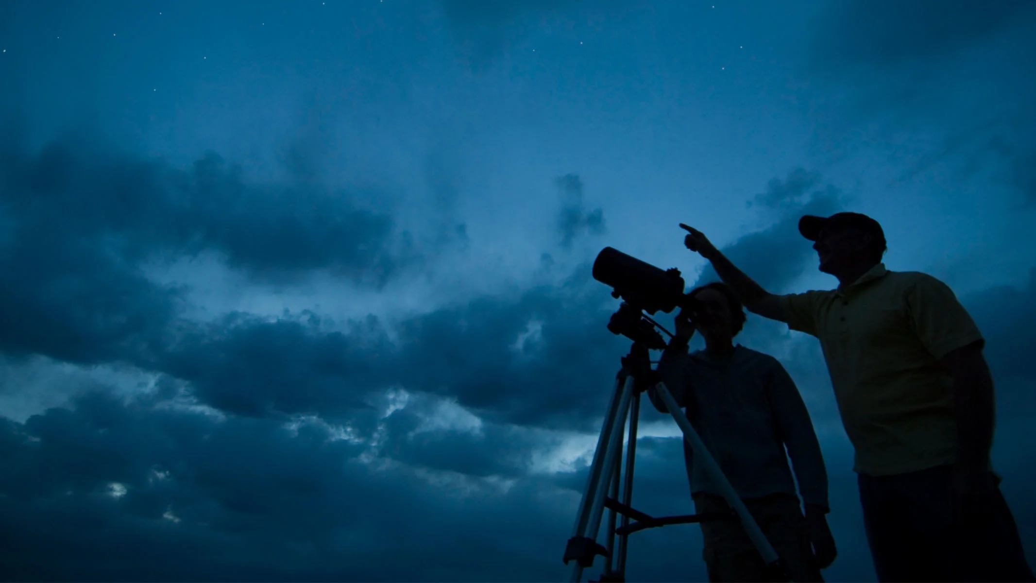 A couple silhouetted against a twilight sky using a telescope.