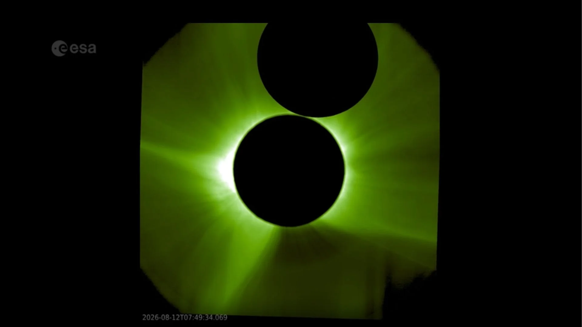 An image of the sun captured by a spacecraft. The sun's corona shines green from behind the black circle of a sunshield, which hides the sun's surface. The moon's silhouette can be seen above.