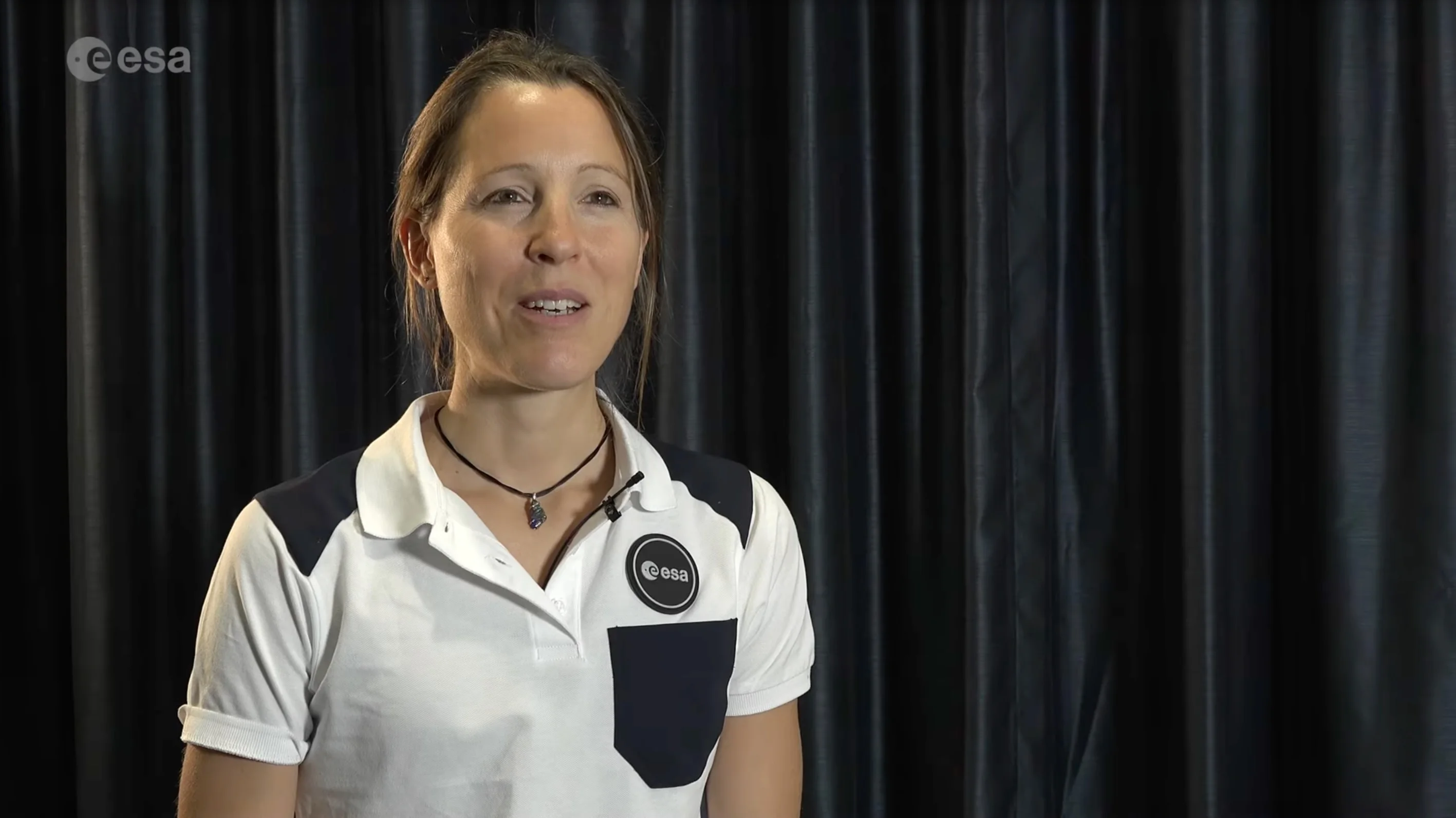 a smiling woman in a white polo shirt sits in front of black drapes