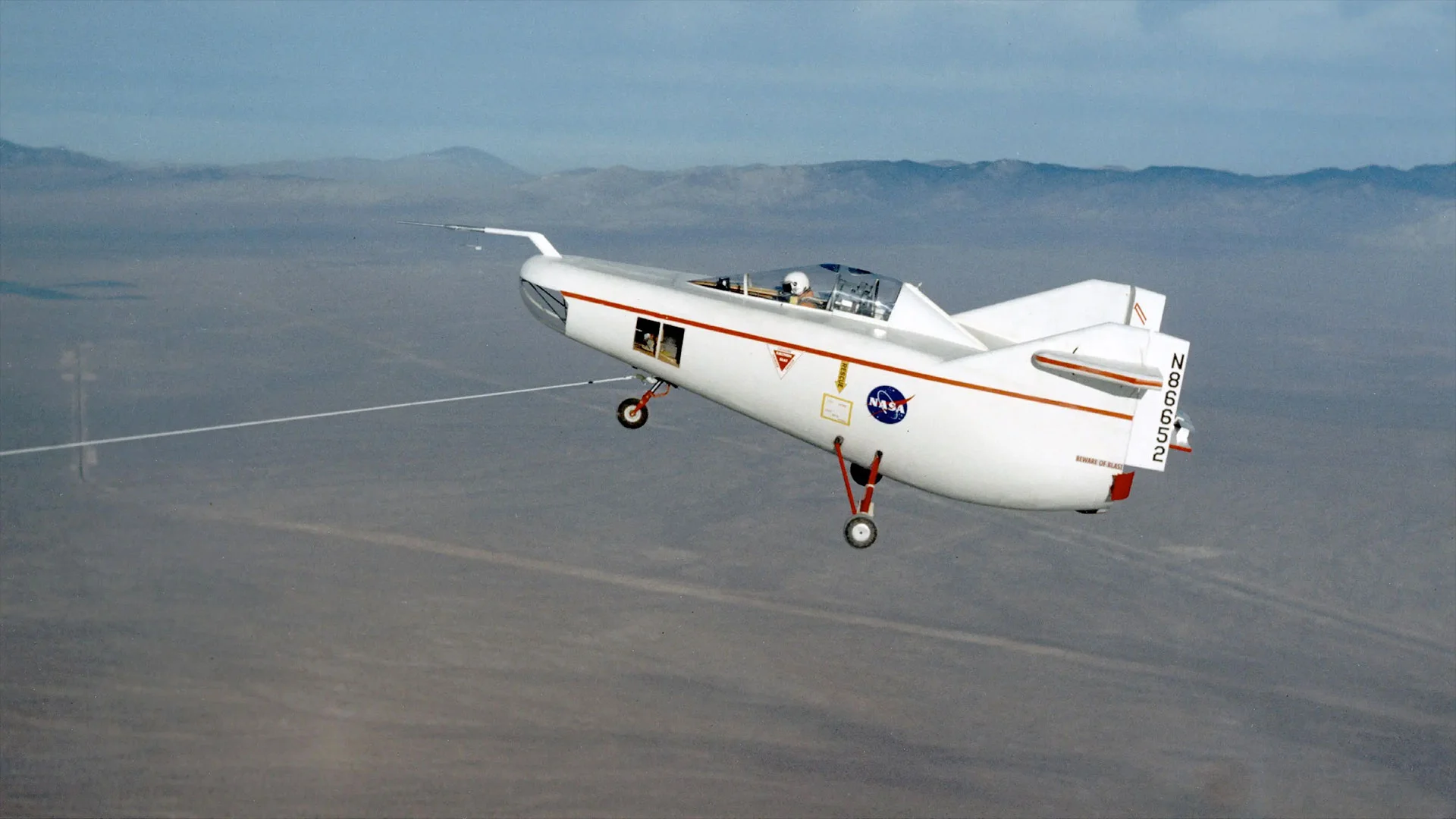 a small white aircraft with a pilot aboard is towed in flight by line extending to the dry lake bed below.