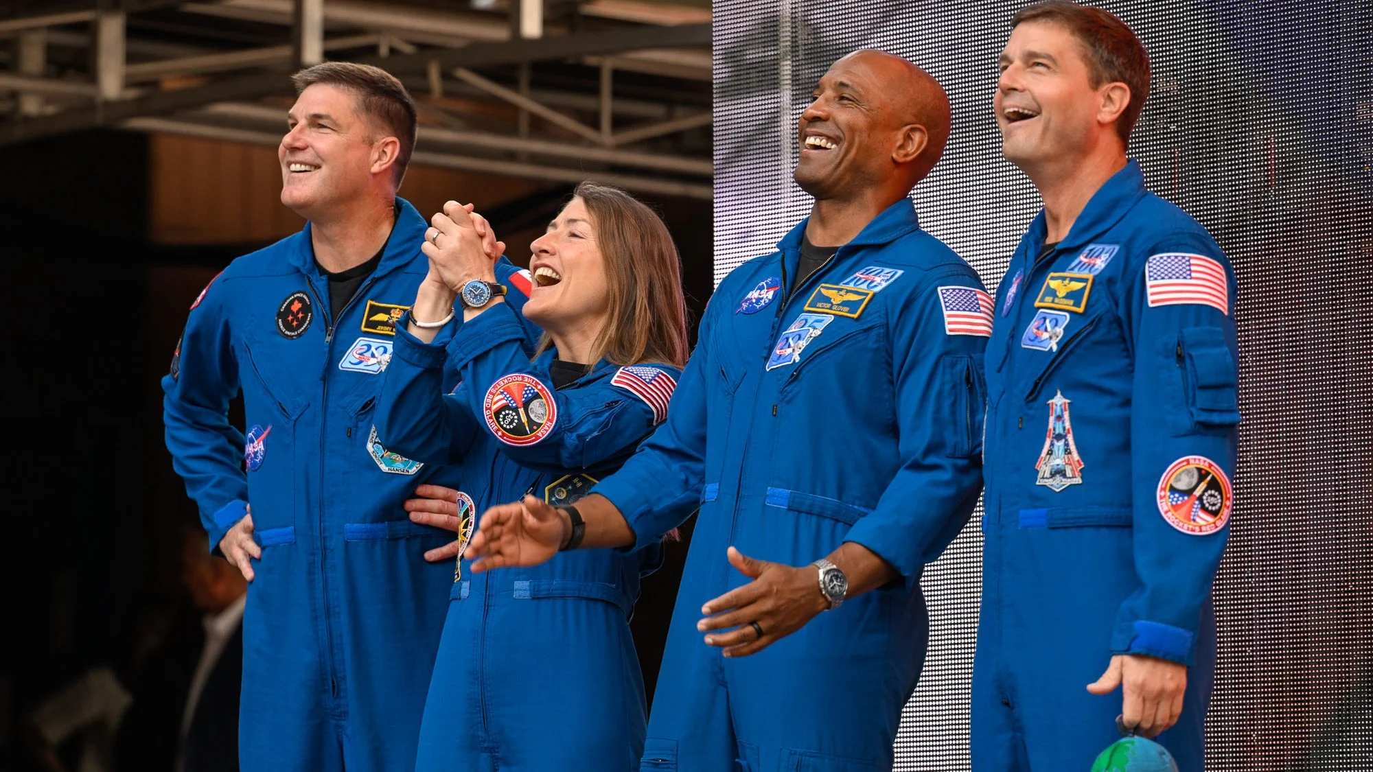 four people in blue flight suits stand on a stage in an amphitheater surrounded by red stone cliffs