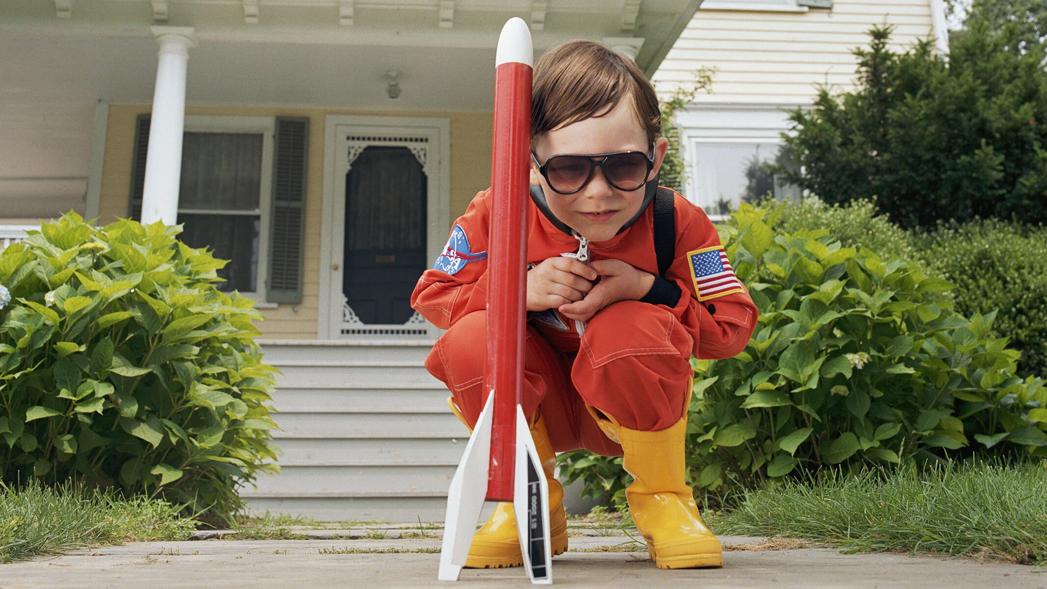 A boy in an astronaut costume crouched down by a toy rocket.