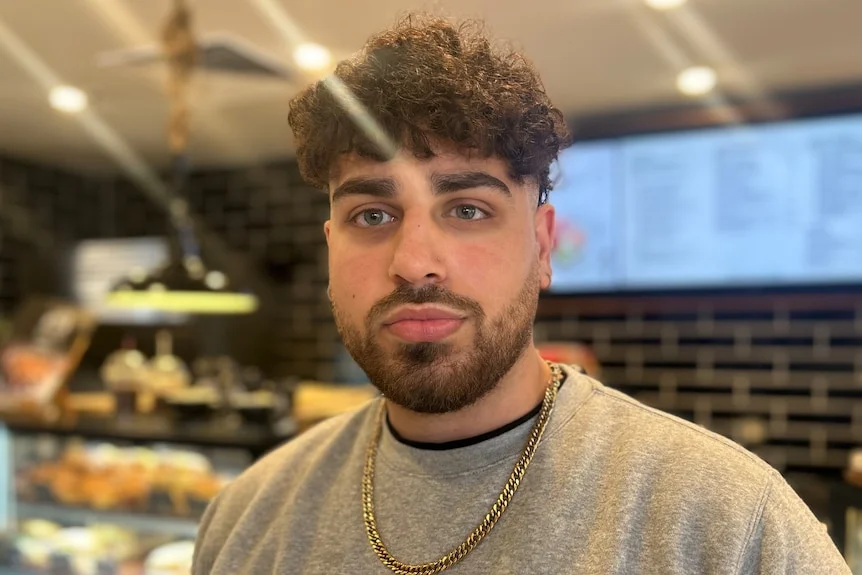 A man with curly brown hair and wearing a grey jumper looks directly at the camera