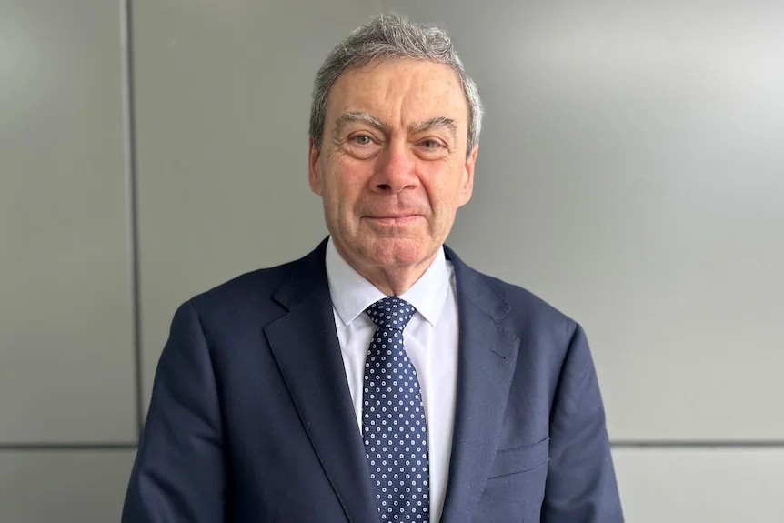 A grey-haired man in a blue suit and tie looks at the camera