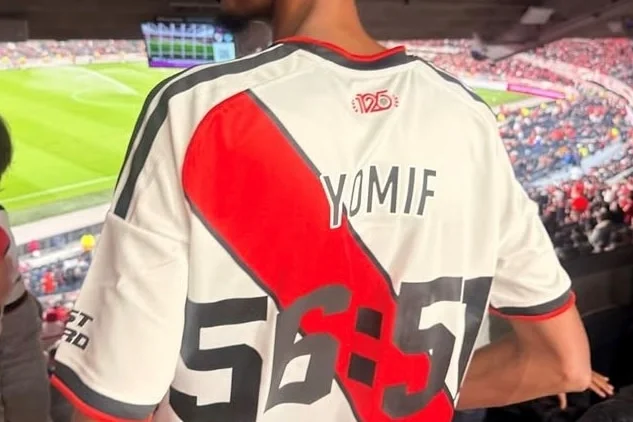 A man in a football jersey watches a match from high in a stadium.