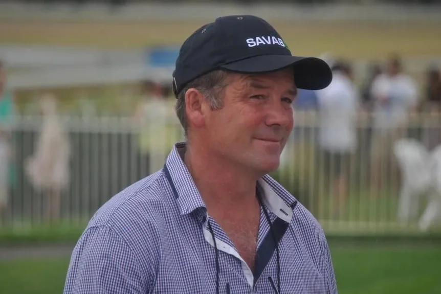 Brett, a smiling man in a cap stands on a racecourse, background blurred. He looks Caucasian.