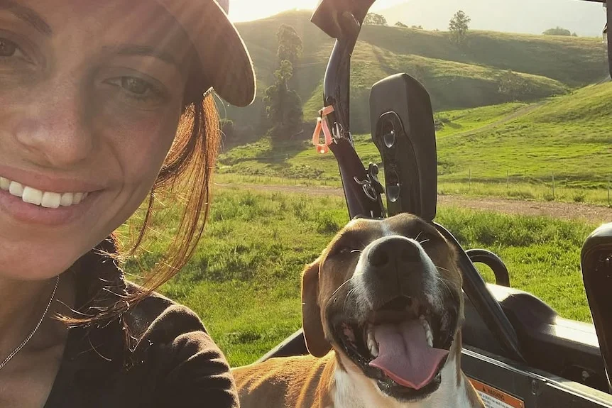 A smiling young woman in a cap takes a selfie with a dog sitting in an ATV on a country property.