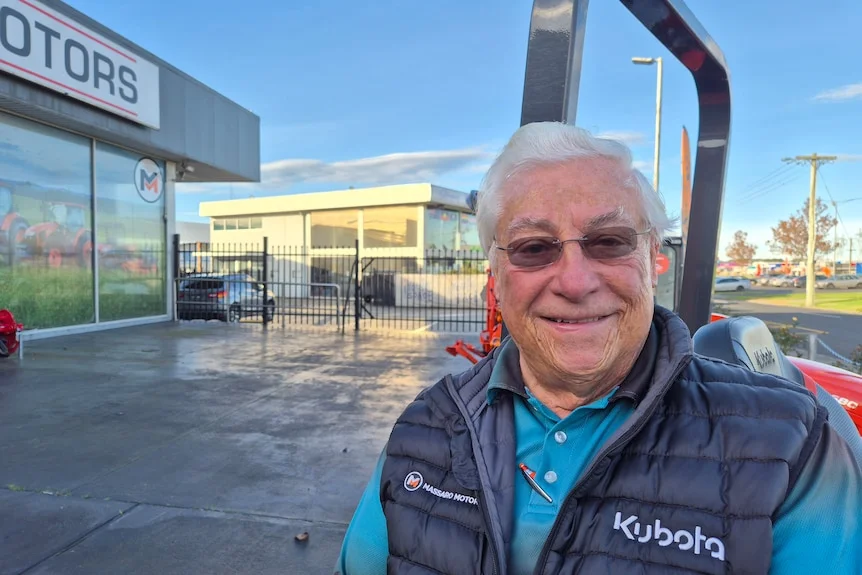 An elderly man in a puffer vest and blue tshirt outside Massaro Motors with Latrobe Youth Space behind him.