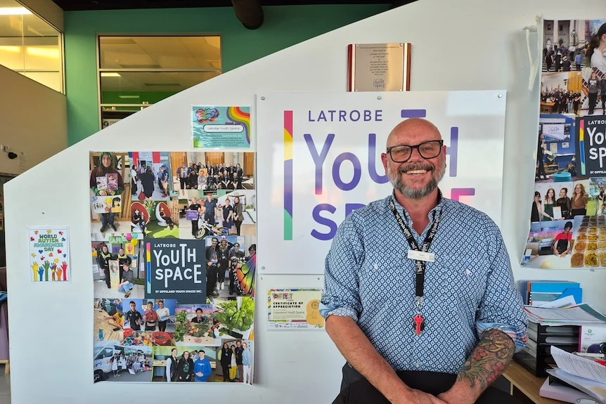 A middle aged man in a business shirt stands in front of Youth Space signs and photos, smiling