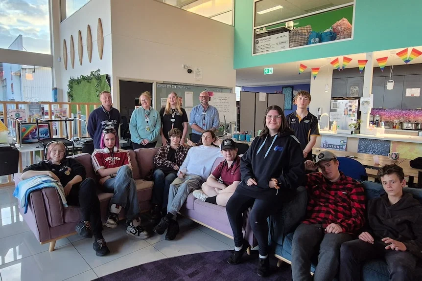 A group of young people in casual clothes sitting on a sofa in a big building, with adults standing behind them.
