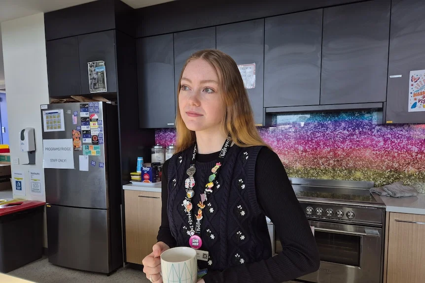A young woman with strawberry blonde hair in a dark top holds a mug in a kitchen, looking thoughtful