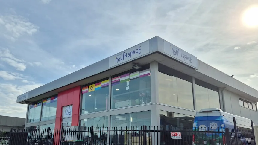A grey building with a red doorway, with the sun behind it and a blue bus next to it.