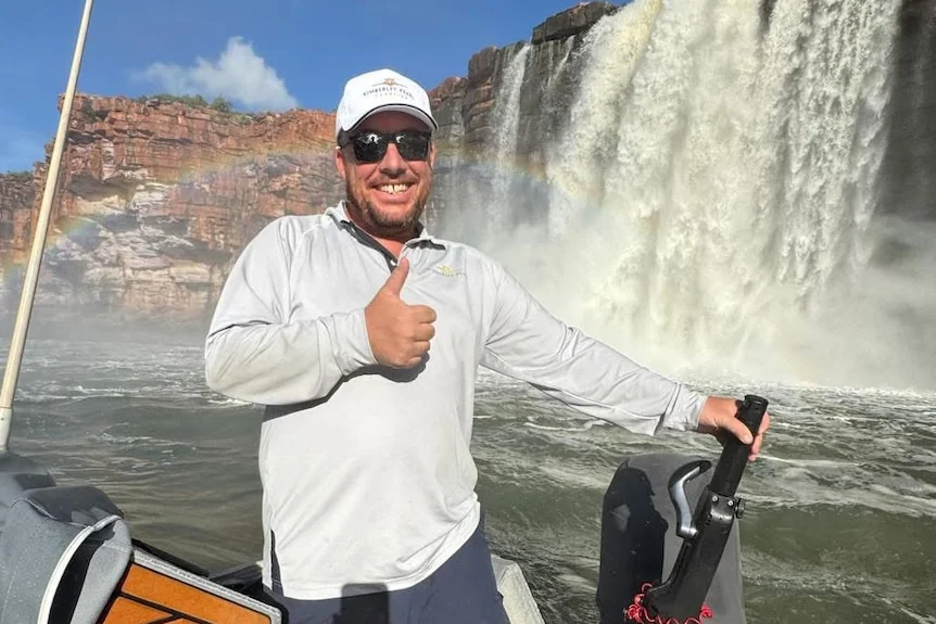 Daniel Brown stands on a boat smiling while giving a thumbs up