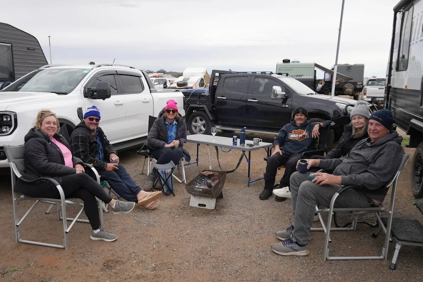 Group of six people sit around campfire on fold up chairs facing camera, next to a table, utes and a caravan