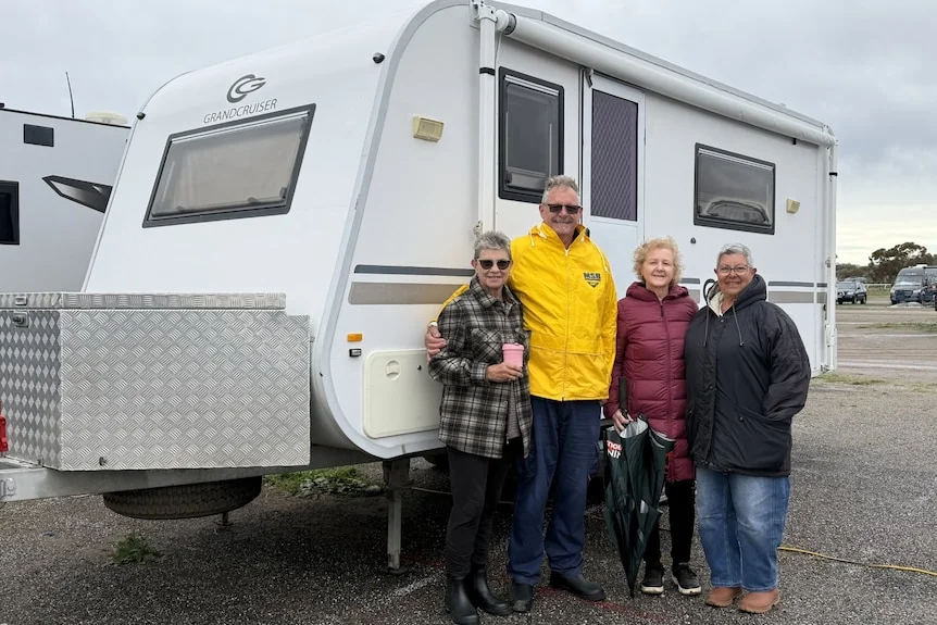 Three women and a man stand in front of a caravan. 