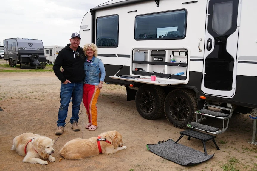 Man and woman stand next to white caravan with two golden retriever dogs in front of them on ground. 