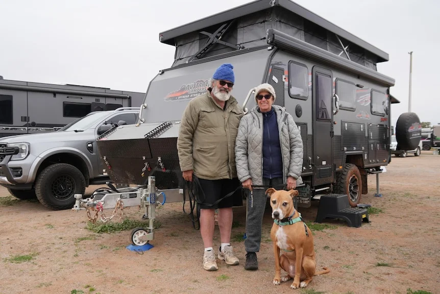 Man and woman stand in front of a caravan with a dog 
