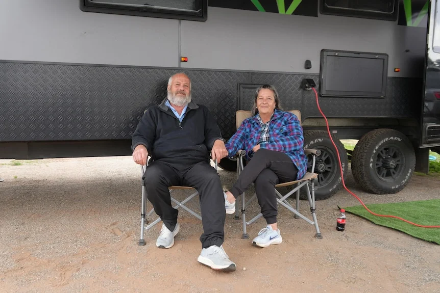 Couple sits on fold up chairs in front of caravan. 