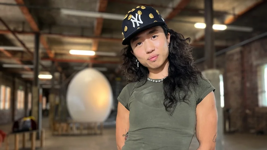 artist EJ Son stands inside a warehouse with a ginat egg inthe background as they look into the camera