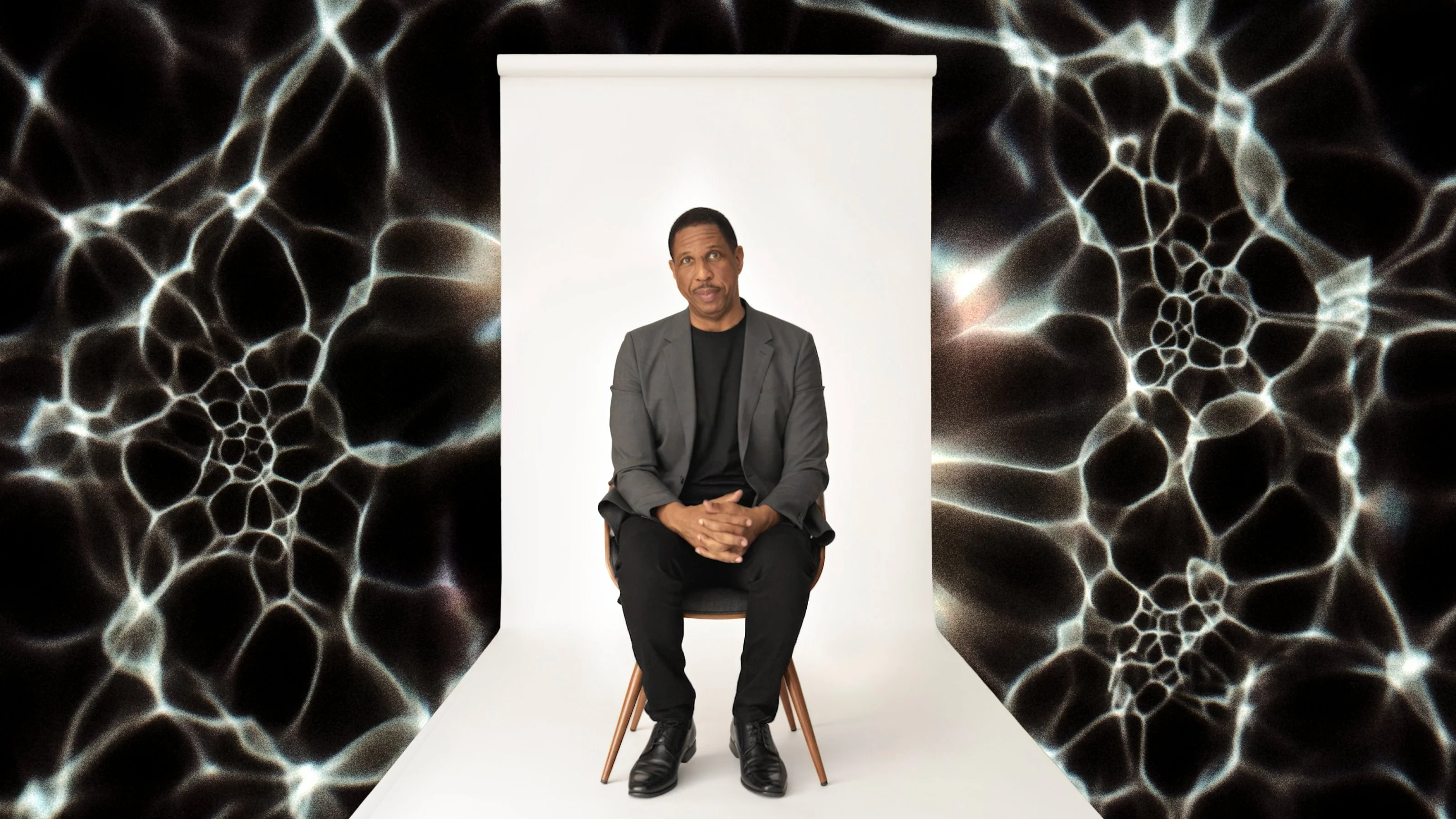 A man in a dark suit sits on a chair against a white backdrop, with abstract black and white patterns surrounding him.
