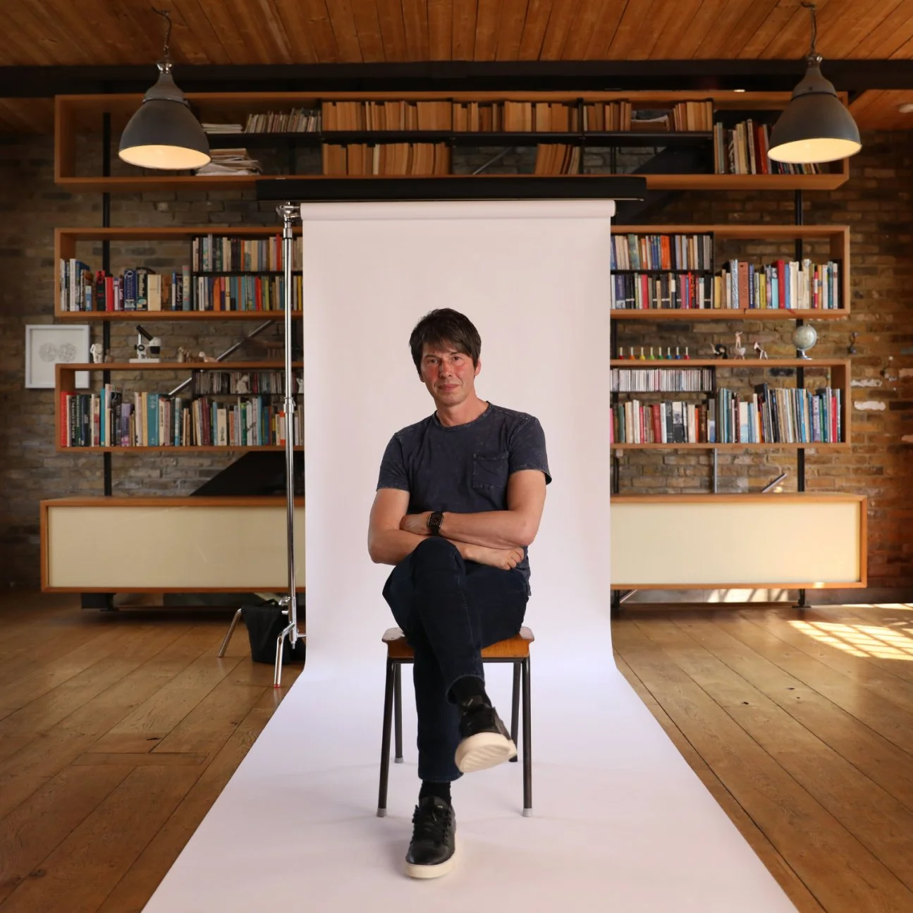 A man in a blue t-shirt and jeans sits on a chair with legs crossed, in front of a white backdrop, with a bookshelf and wooden flooring in the background.