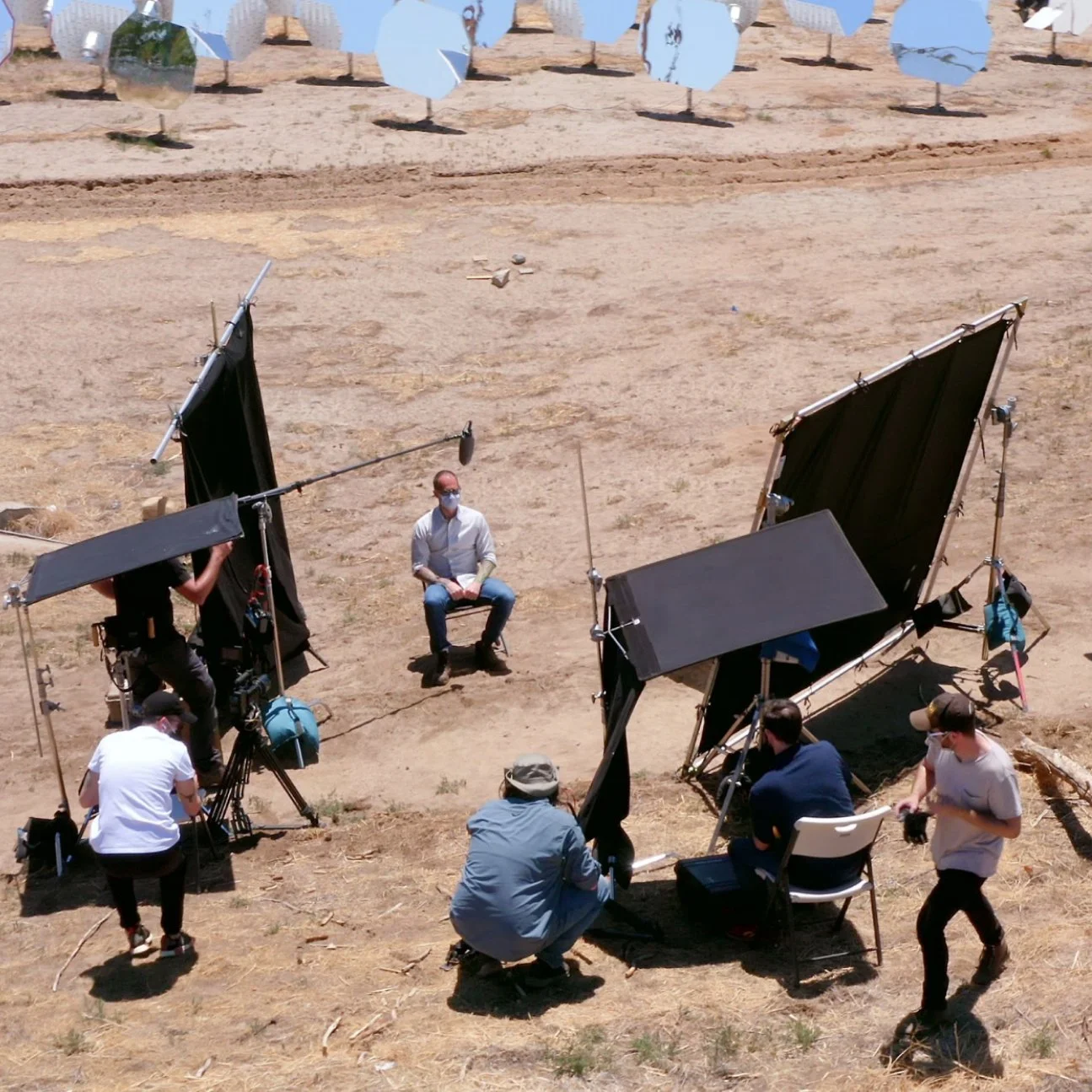 A man sits for an outdoor interview surrounded by cameras, crew, and light reflectors on a dry, grassy field.