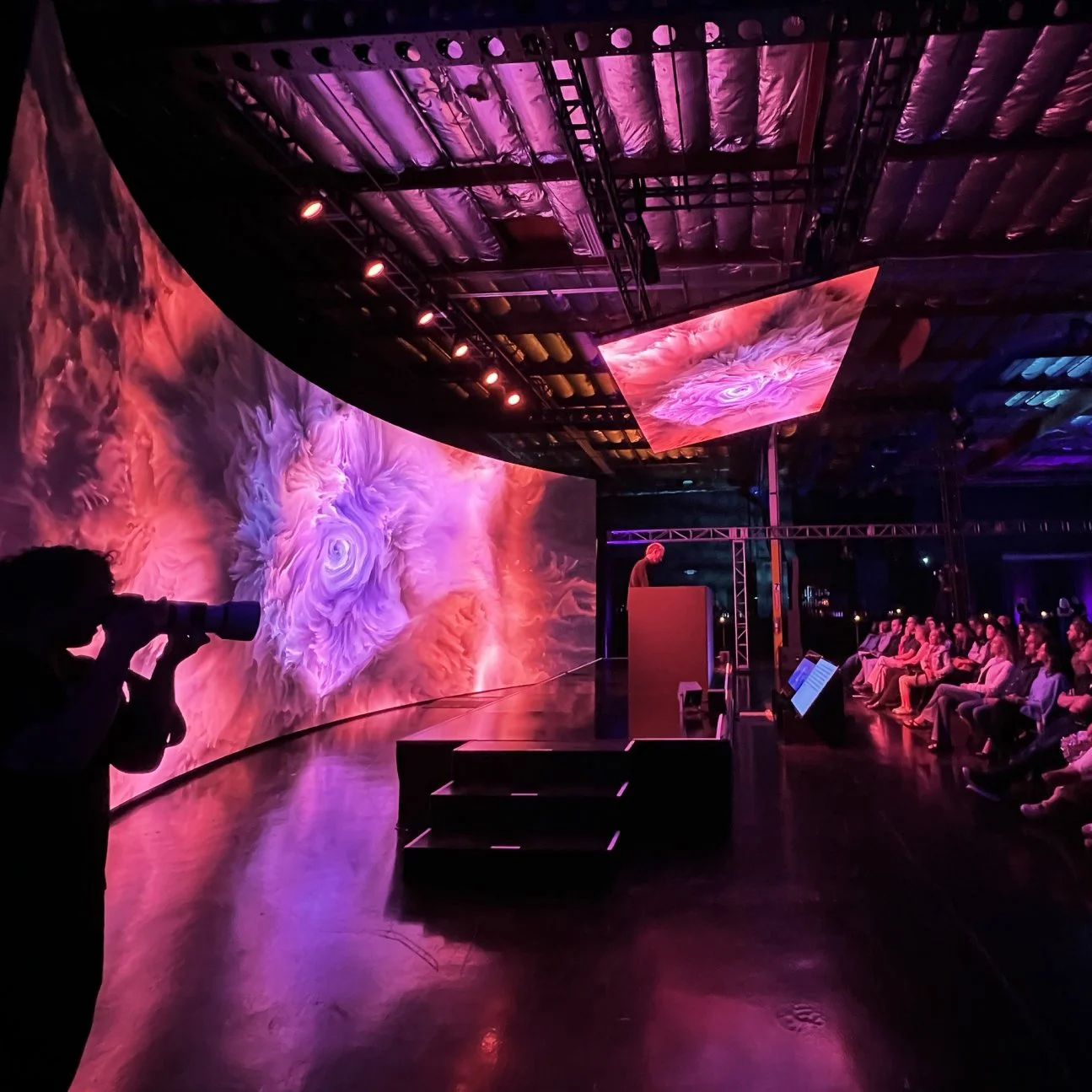 A speaker stands at a podium on a stage with abstract visuals projected behind them, as an audience watches and a photographer captures the scene.