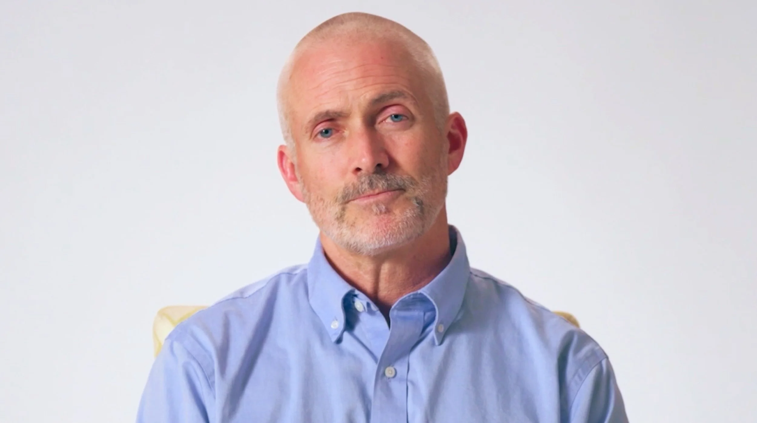 A middle-aged man with a shaved head and trimmed beard, wearing a light blue button-down shirt, looks directly at the camera against a plain light background.