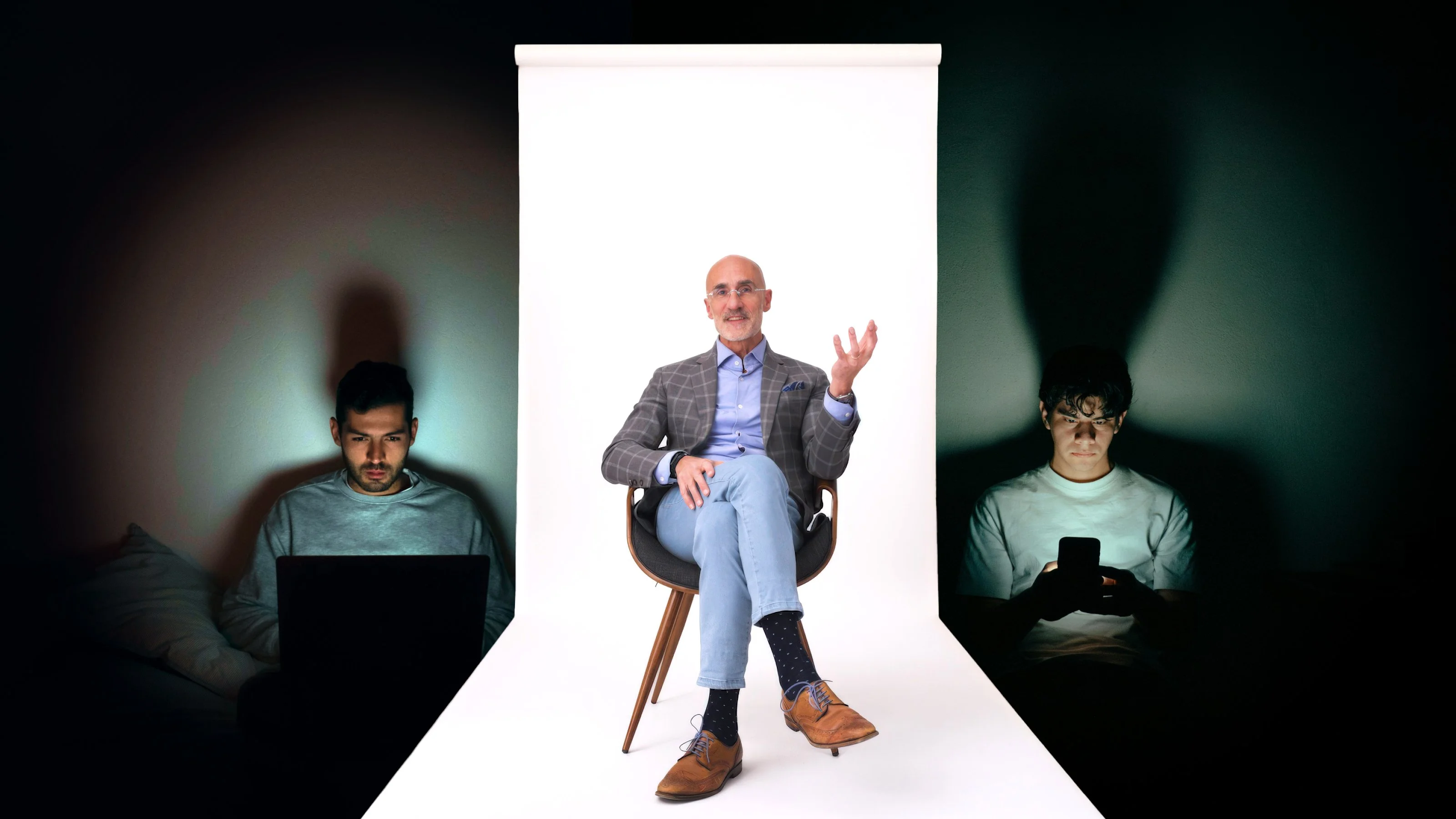 A man in a suit sits on a chair in front of a white backdrop, flanked by two people using electronic devices in dim lighting.