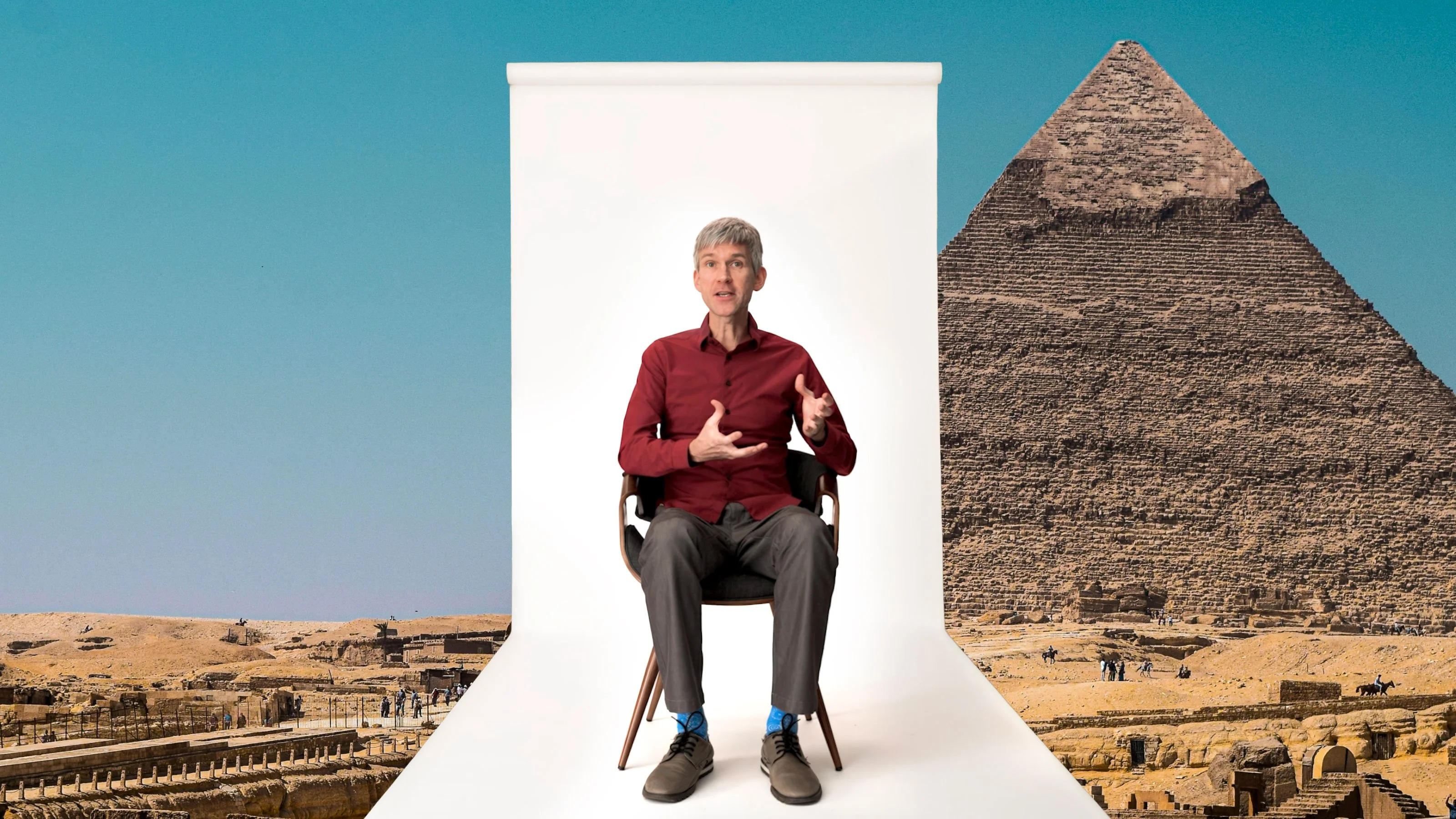 A man sits on a chair against a white backdrop, placed in front of the Pyramid of Giza in Egypt, under a clear blue sky.