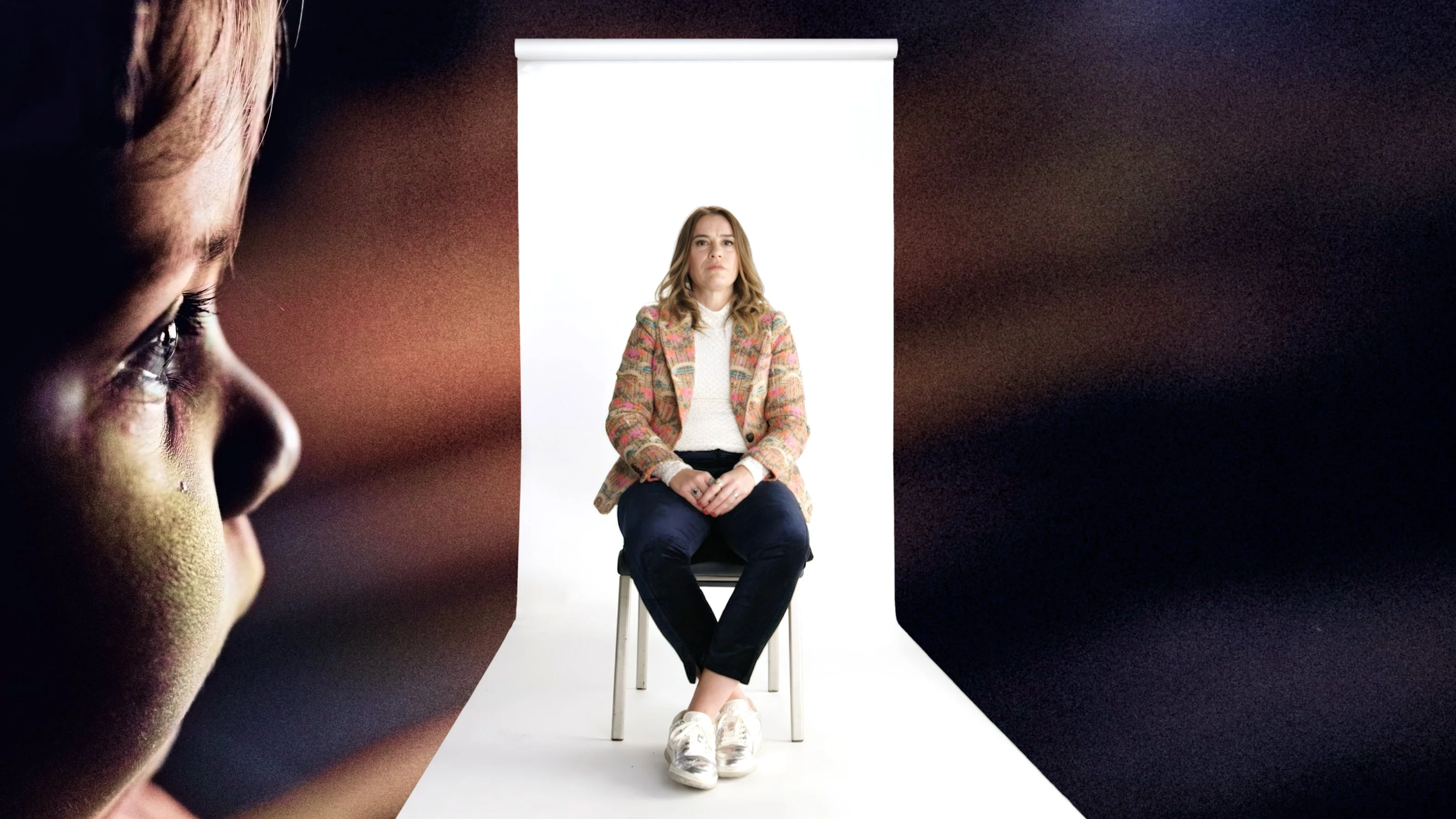 A woman sits on a chair in front of a white backdrop; the blurred profile of a child with a tear rolling down their cheek is visible in the foreground.