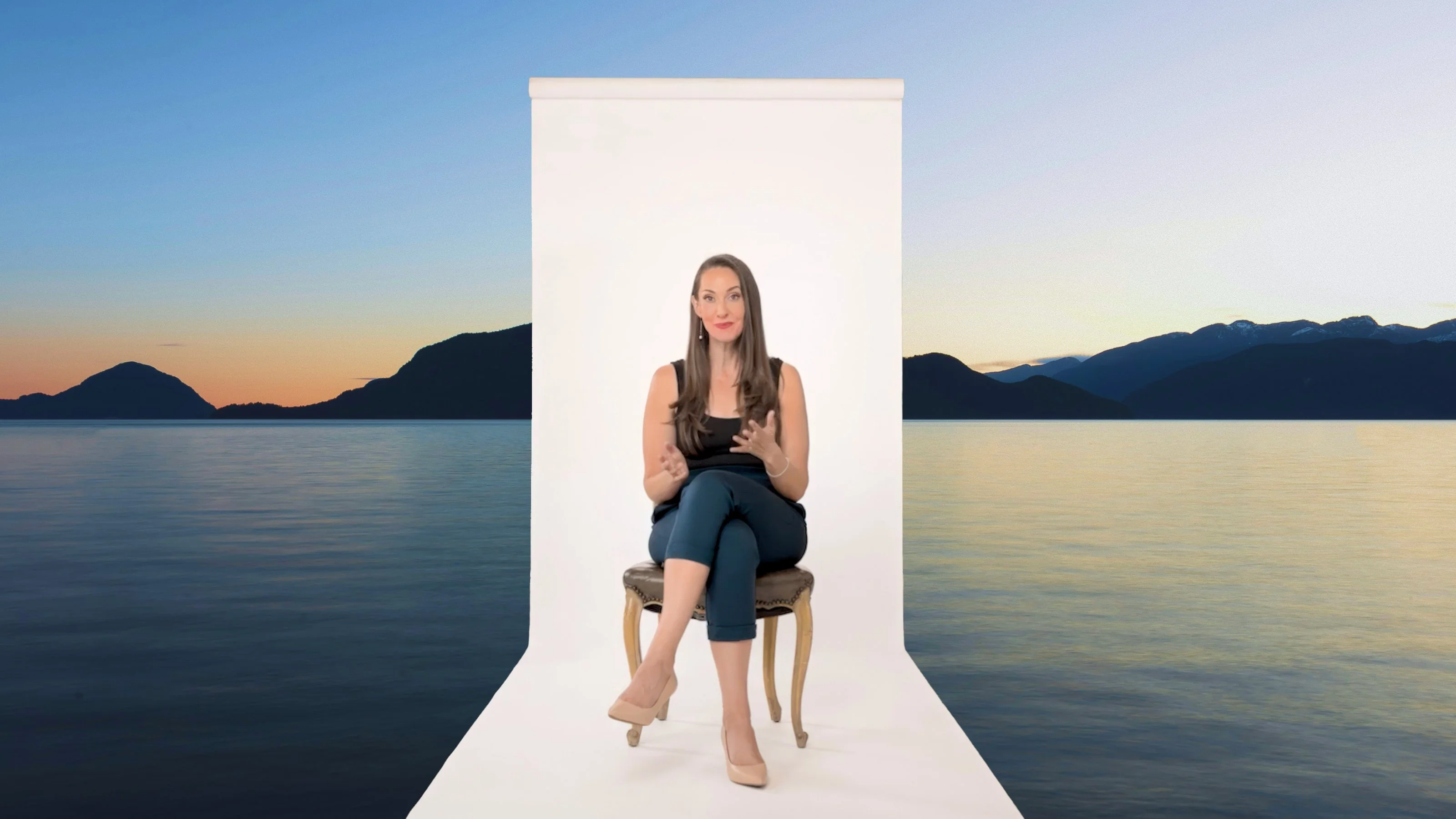 A woman sits on a chair in front of a white backdrop set against a scenic lake and mountain landscape at sunset.