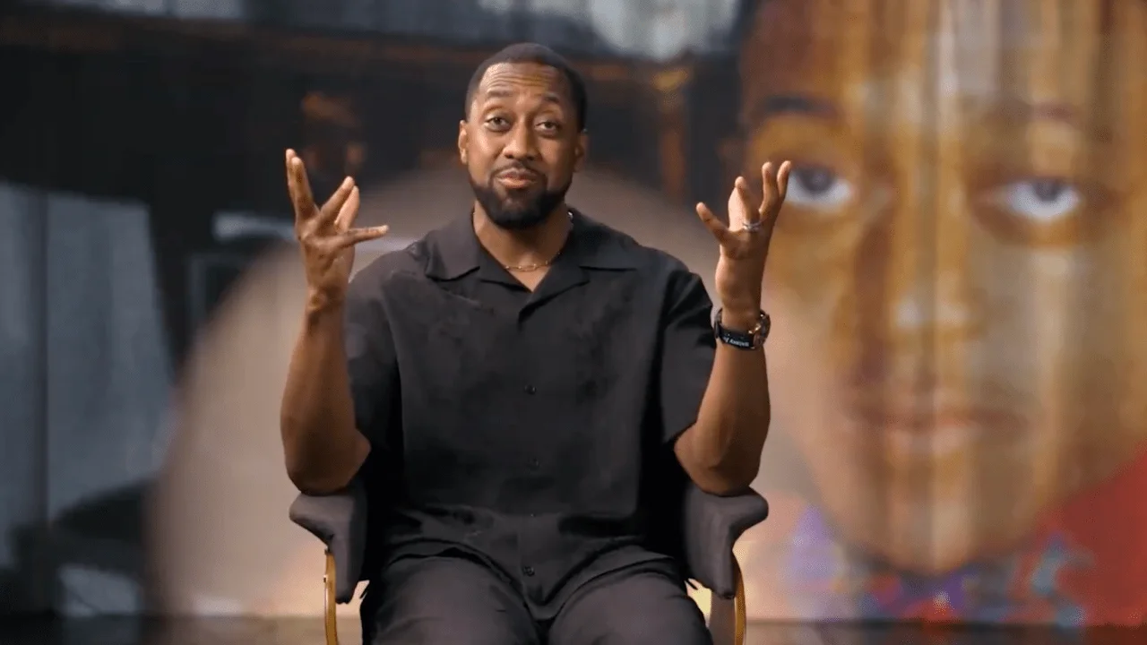A man in a black shirt sits on a chair, gesturing with his hands, with a large blurred image of his younger self in the background.