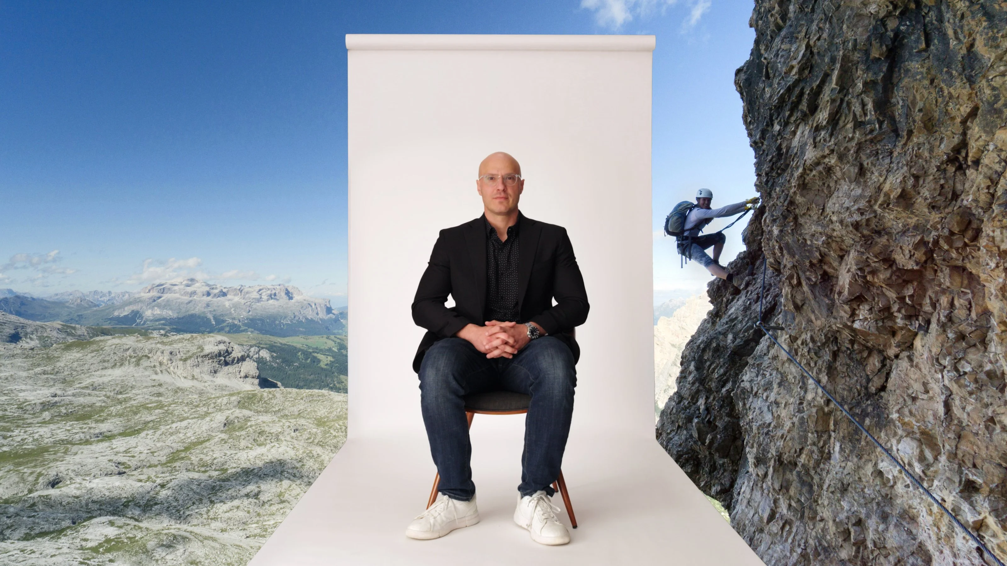A man in casual business attire sits on a chair in front of a white backdrop, with a mountain landscape and a climber scaling a rocky cliff in the background.