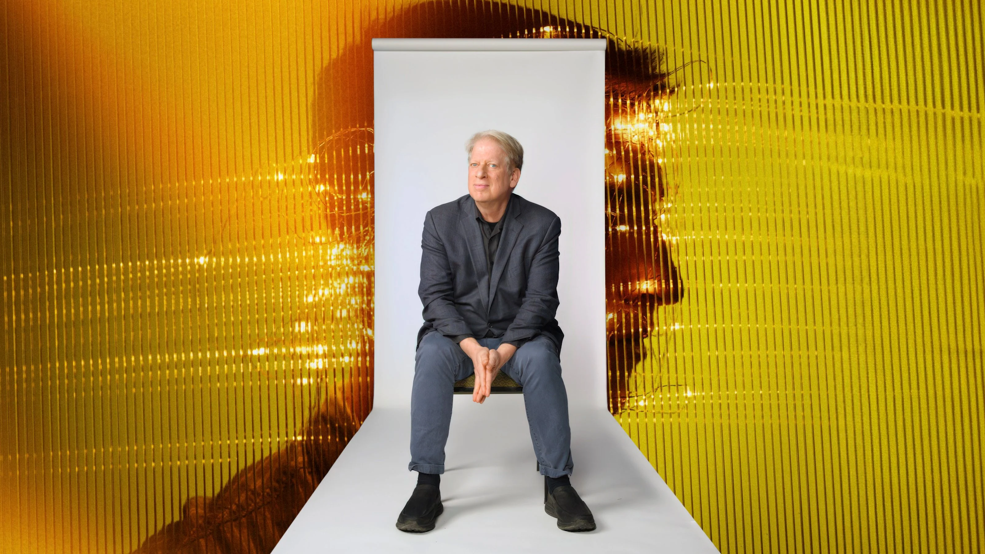 A man sits on a stool against a small white backdrop, with a large yellow silhouette and abstract pattern in the background.