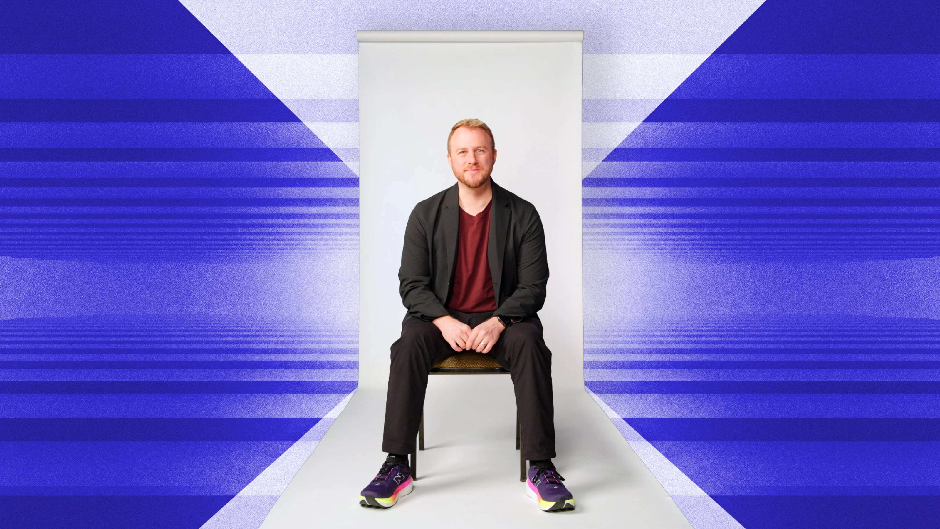 A man with light hair sits on a chair in front of a white backdrop, with a geometric blue and white pattern in the background.