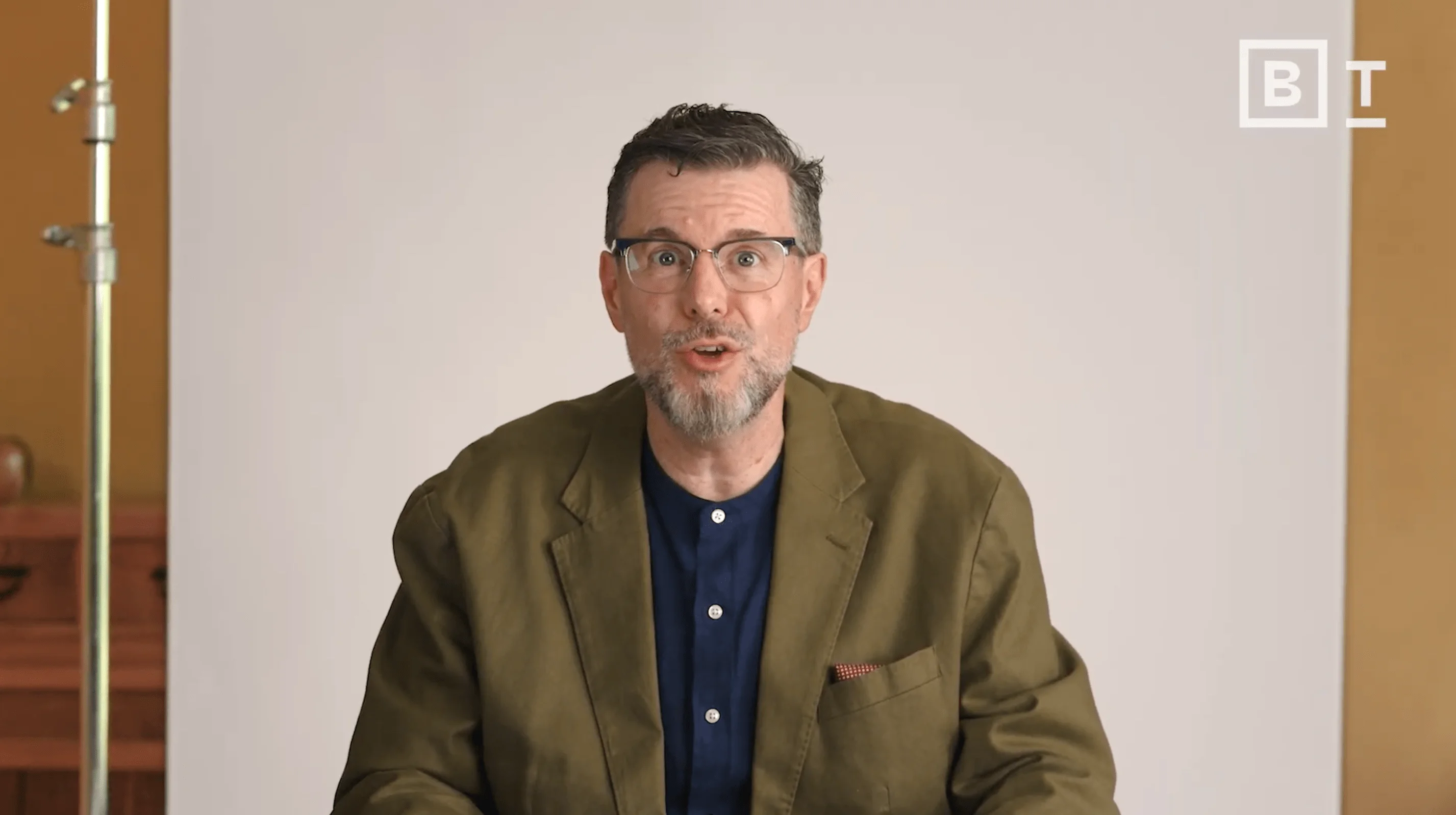 A man with glasses and a beard wearing a green blazer and blue shirt sits in front of a plain backdrop, looking at the camera. The letters "BT" are visible in the top right corner.